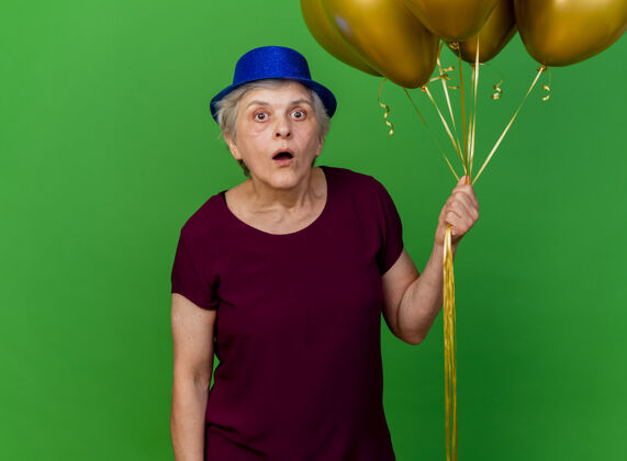 氦戴着派对帽的震惊的老妇人拿着绿色的氦气球派对老人帽子