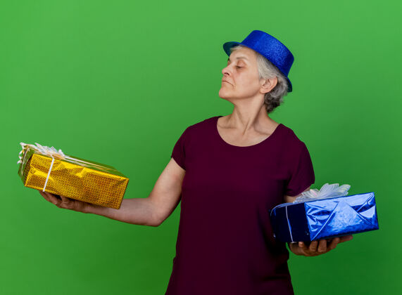 老人戴着派对帽的自信的老妇人拿着绿色的礼品盒看盒子礼物自信