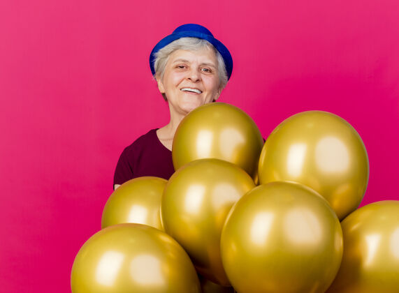 女人微笑的老妇人戴着派对帽站在氦气球上看着粉红色的相机帽子粉色老人