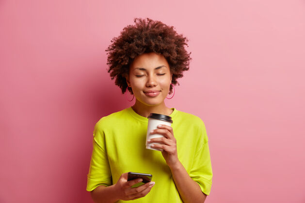 网络快乐的年轻卷曲的非洲裔美国女人闭上眼睛闻着令人愉快的咖啡香味有着放松的脸用智能手机上网聊天穿着休闲装隔离在粉红色的墙上交流年轻欢呼