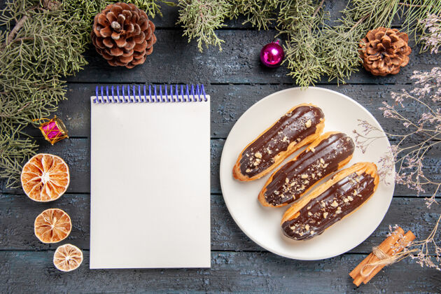 桌子俯视图白色椭圆形平板上的巧克力eclairs冷杉树枝和圆锥体圣诞玩具干橘子肉桂和一个笔记本在深色的木桌上食物午餐干的