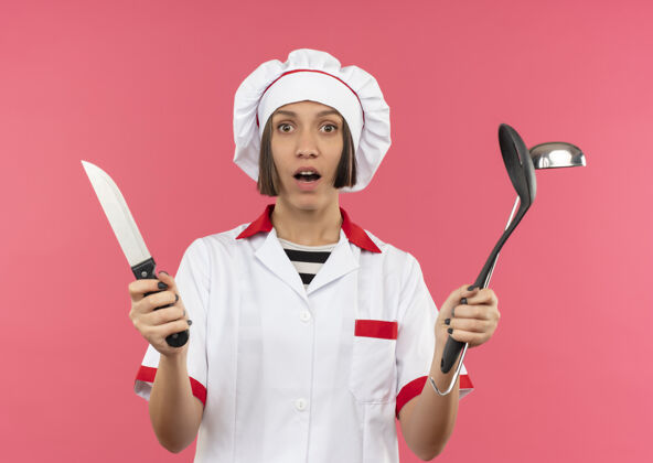 姿势惊讶的年轻女厨师穿着厨师制服拿着刀 抹刀和勺子隔离在粉红色的墙上女性烹饪惊喜