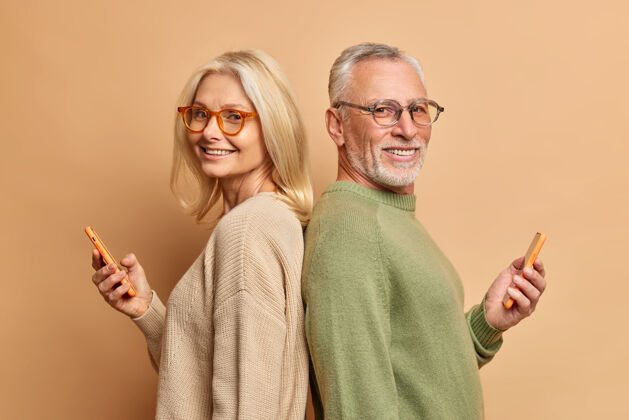 快乐高级男女背靠背站立使用现代手机背靠背站立戴眼镜和休闲毛衣隔着棕色的墙壁年龄爷爷丈夫