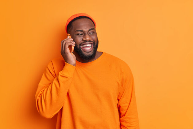 帅气开朗的胡须男人打电话时笑容满面 牙齿洁白 穿着橙色毛衣 戴着帽子 高兴地一边看一边讨论周末的计划科技时髦单色