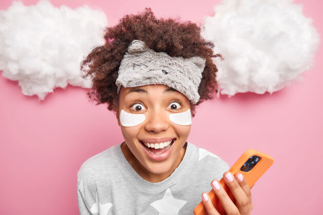 成人一个穿着睡衣 戴着口罩的快乐的美国黑人妇女用手机在镜头前愉快地看着镜头 为她的博客写了一篇文章 眼睛下面贴着胶原蛋白 在云端摆出姿势人休息头发