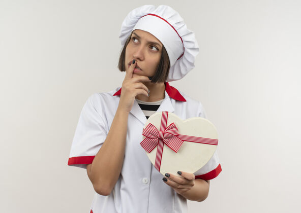人体贴的年轻女厨师 身着厨师制服 手持心形礼盒 手指放在嘴唇上 看着隔离在白色墙上的一边人烹饪感情