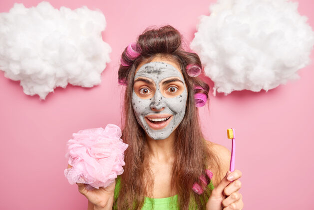 快乐正面黑发女人用发卷做发型拿淋浴海绵刷牙用刷子在脸上涂有滋养泥面膜隔离粉色墙壁健康美女女人
