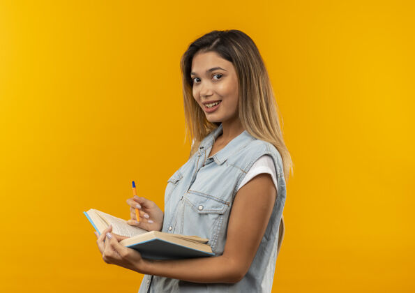 抱微笑着的年轻漂亮的学生女孩 背着书包站在侧视图中 手里拿着打开的书和笔 孤立地站在橙色的墙上脸衣服包