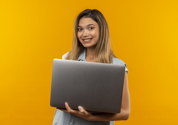 漂亮微笑着的年轻漂亮的学生女孩穿着背包拿着笔记本电脑孤立在橙色的墙上衣服表情人