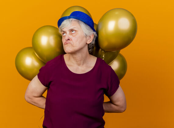 悲伤戴着派对帽的悲伤的老妇人在身后举着氦气球仰望橙色气球老人穿