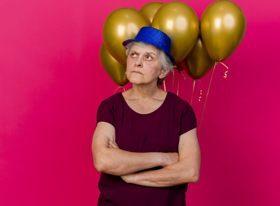 女人戴着派对帽的严肃的老妇人站在氦气球前 双臂交叉在粉红色的气球上帽子严肃氦