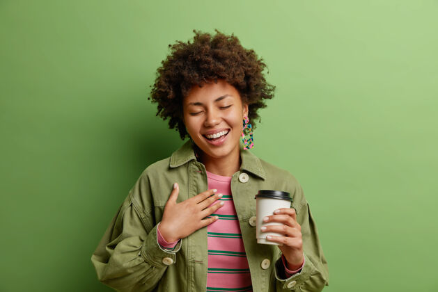 高兴积极的非裔美国女性 卷发闭上眼睛远离幸福 手靠近心脏 喝外卖咖啡 穿着时髦的衣服 享受休闲时光 隔离在绿色的墙壁上饮酒真诚黑发