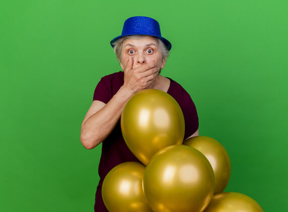 老人震惊的老妇人戴着聚会帽 手放在嘴上 站在绿色的氦气球上派对氦手