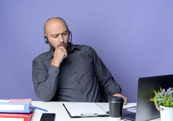 手体贴的年轻秃头呼叫中心男子戴着耳机坐在办公桌旁 手放在下巴上 手放在紫色的墙上 看着前面的工作工具工作人桌子