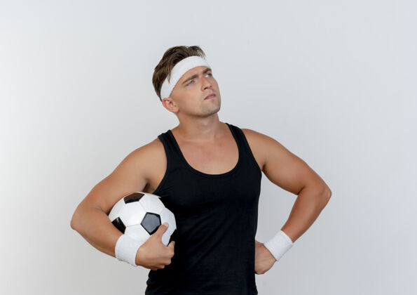 腰体贴的年轻英俊的运动型男子戴着头带和腕带拿着足球 手放在腰上看着隔离在白墙上的一面人姿势抱着
