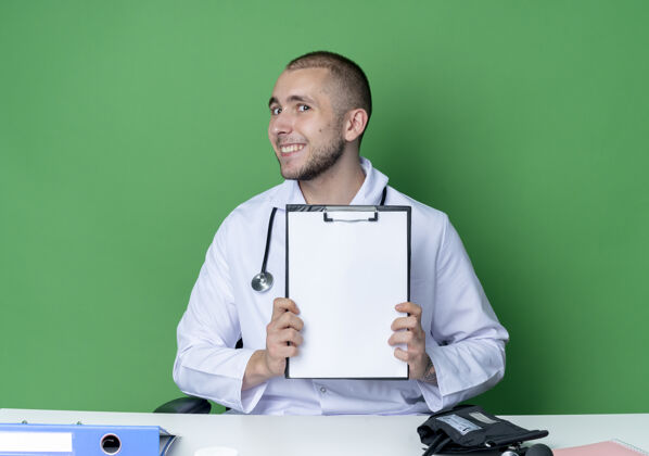 人面带微笑的年轻男医生穿着医用长袍 听诊器坐在办公桌旁 工作工具上的剪贴板被隔离在绿色的墙上感情衣服桌子