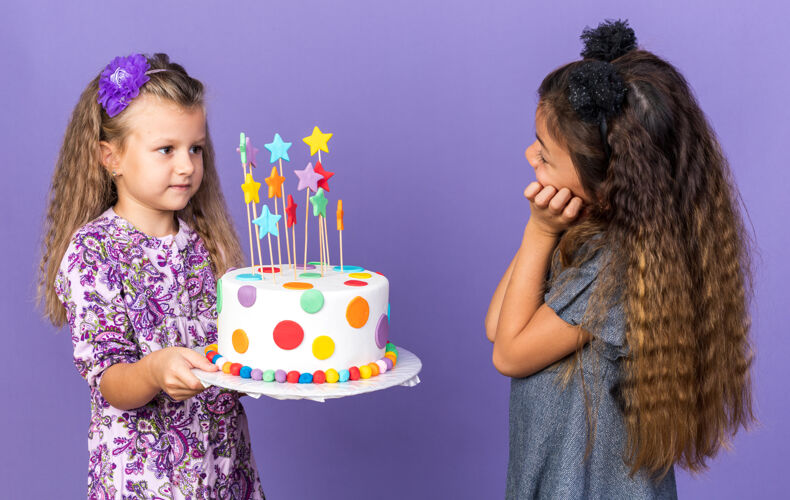 蛋糕高兴的小白种女孩手牵手在一起 看着金发小女孩拿着生日蛋糕孤立在紫色墙上的复制空间孤立女孩小