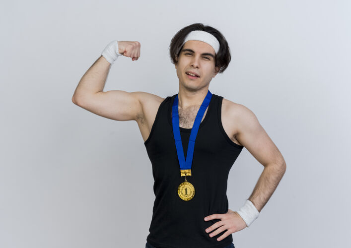 信心年轻的运动型男子身穿运动服 头戴金牌 脖子上举着拳头 站在白色的墙上 二头肌看起来很自信脸姿势提高