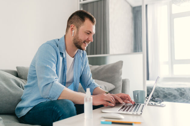 计算机穿着衬衫的微笑男人在家里的沙发上轻松地坐在桌子旁 在笔记本电脑上在线工作坐姿人室内