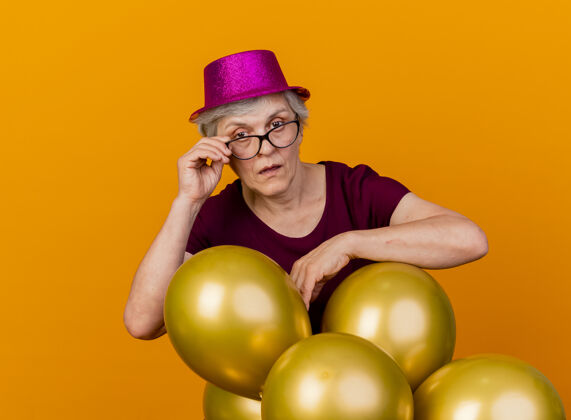 气球自信的老妇人戴着派对帽站在那里 手里拿着氦气球 透过隔离在橙色墙上的光学眼镜看着前方人党姿势