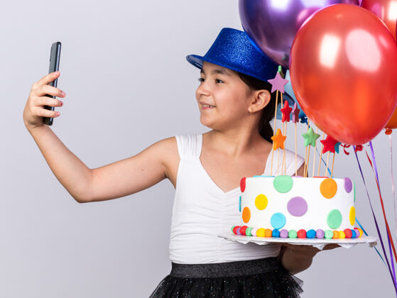 女孩带着蓝色派对帽 手持氦气球和生日蛋糕的年轻高加索女孩微笑着在手机上自拍 被隔离在白色墙壁上 留有复印空间帽子气球采取