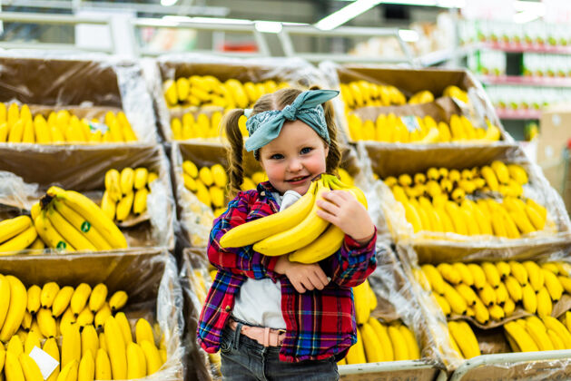 小女孩可爱的小女孩拿着香蕉在食品店或超市买水果快乐