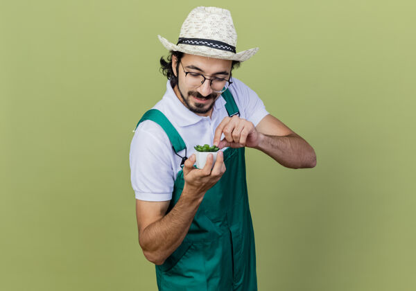 光年轻的留着胡须的园丁 穿着连体衣 戴着帽子 看着盆栽植物 站在浅绿色的墙上微笑着看着它站穿脸
