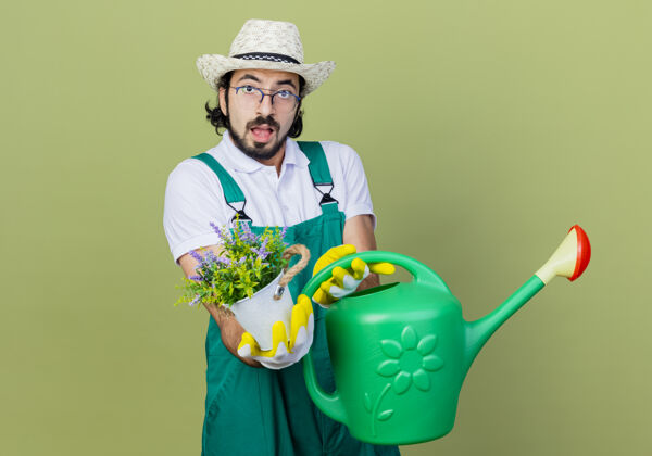 帽子年轻的留着胡须的园丁 穿着连体衣 戴着帽子 拿着水罐和盆栽植物 站在浅绿色的墙上 困惑地看着前方浇水年轻连身衣