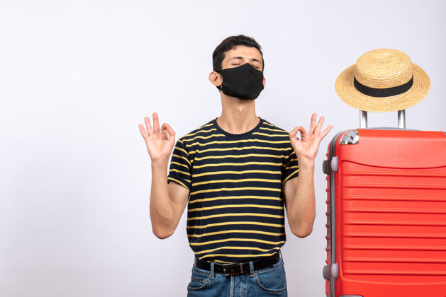 手提箱正面图：戴着黑色面具的年轻游客站在红色手提箱旁 打着“OK”的手势红男前面