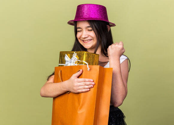 白种人兴奋的年轻白人女孩 戴着紫色派对帽 拿着购物袋里的礼盒 在橄榄绿的墙壁上举起拳头 留着复制空间孤立女孩聚会