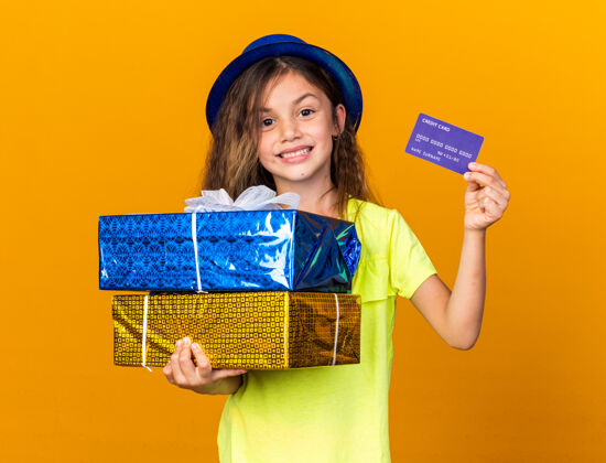 小微笑的白人小女孩 戴着蓝色派对帽 手里拿着信用卡和礼品盒 隔离在橙色的墙上 还有复印空间举行盒子聚会