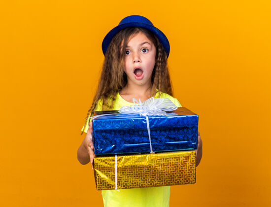 盒子兴奋的小白种女孩 戴着蓝色派对帽 拿着礼品盒 隔离在橙色的墙上 还有复印空间抱着孤立帽子