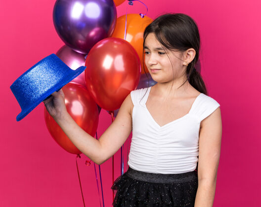 不愉快一个年轻的白人女孩拿着蓝色的派对帽 看着站在粉色墙壁上的氦气球前面 上面有复制空间白种人气球抱着