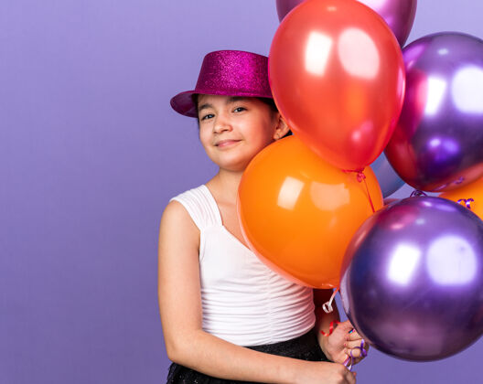 帽子年轻的白人女孩戴着紫色的派对帽 手里拿着氦气球 在紫色的墙壁上隔离着复制空间抱着的氦女孩