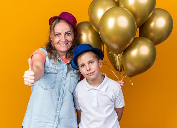 拇指戴着蓝色派对帽的年轻斯拉夫男孩高兴地站在那里 母亲戴着紫色派对帽 手里拿着氦气球 竖起大拇指 孤立地站在橙色的墙上 留着复制空间站着孤立的男孩
