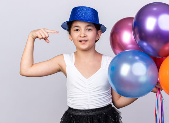 指着戴着蓝色派对帽的年轻高加索女孩高兴地拿着 指着隔离在白色墙壁上的氦气球 还有复印空间白种人孤立的拿着