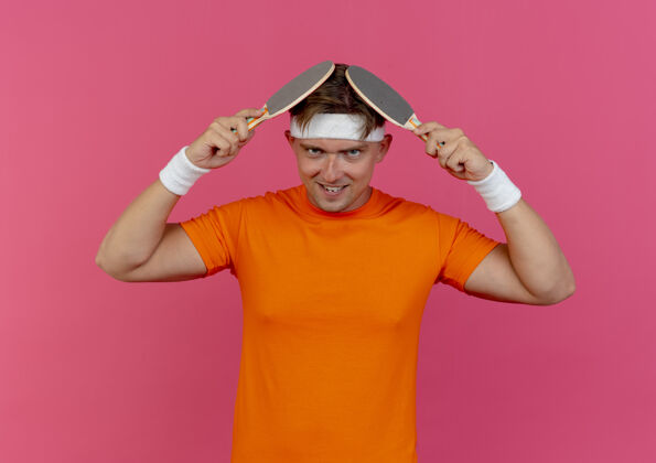 头带面带微笑的年轻帅气的运动型男子戴着头带和腕带 用隔离在粉红色墙上的乒乓球拍碰头表情触摸头