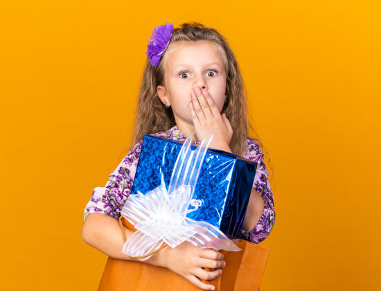 女孩惊讶的小金发女孩把手放在嘴上 拿着礼品盒孤立地放在橙色的墙上复制空间金发惊喜孤立