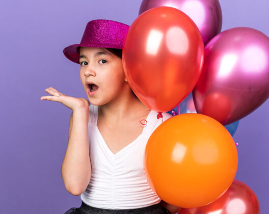 年轻惊讶的年轻白人女孩戴着紫色的派对帽 手里拿着氦气球 手一直张开 在紫色的墙上留着复制空间手女孩孤立