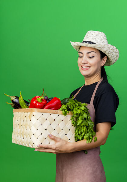美丽年轻漂亮的女园丁 围着围裙 戴着帽子 手里拿着装满蔬菜的篮子 站在绿色的墙上 满脸笑容女人帽子脸