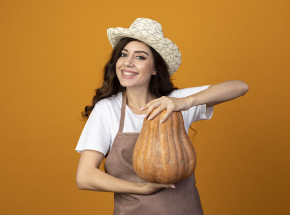 表情身着制服 头戴园艺帽 面带微笑的年轻女园丁把南瓜孤立在橙色的墙上南瓜市民人
