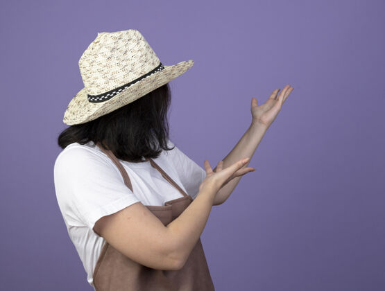表情令人印象深刻的年轻黑发女园丁穿着制服 戴着园艺帽 看着隔离在紫色墙上的一边姿势年轻人脸
