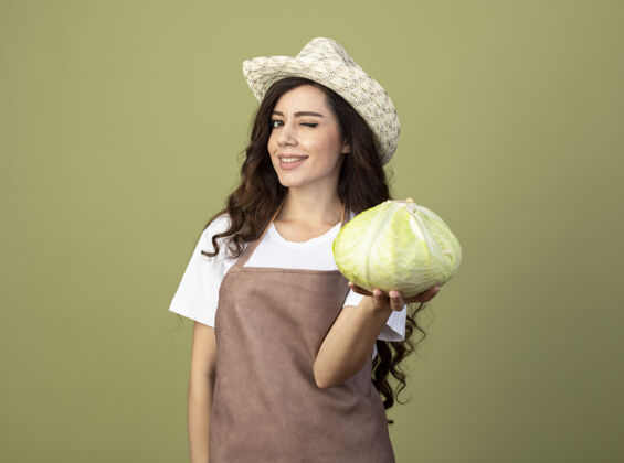 表情自信的年轻女园丁穿着制服 戴着园艺帽 眨着眼睛 把卷心菜隔离在橄榄绿的墙上女人眼睛园艺