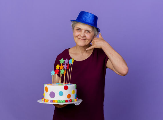 姿势戴着聚会帽的老太太高兴地打着手势叫我 并把生日蛋糕孤立地放在紫色的墙上市民人人