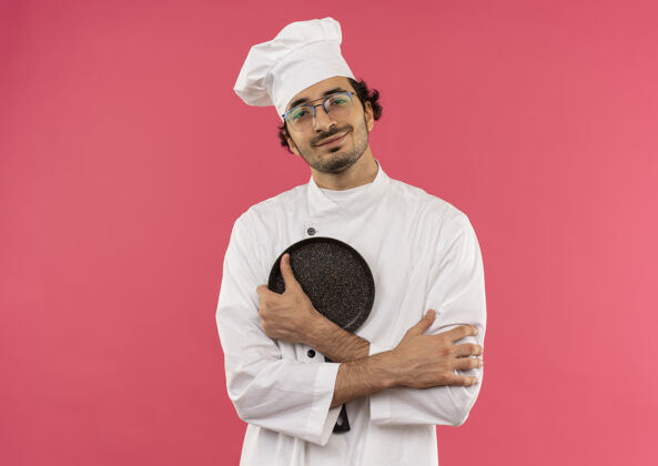 男性面带微笑的年轻男厨师穿着厨师制服 戴着眼镜 双手交叉 手里拿着煎锅粉色交叉微笑