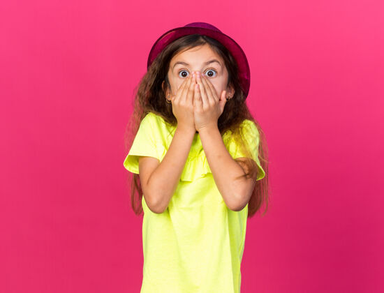小的震惊的小白种女孩 戴着紫色派对帽 手放在嘴上 隔离在粉红色的墙上 留有复制空间孤立的女孩白种人