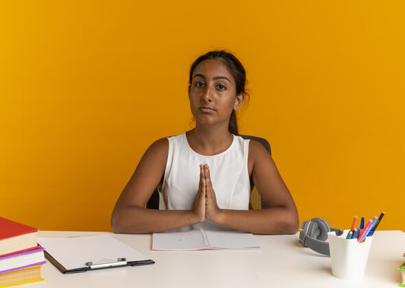 祈祷看着镜头 年轻的女学生坐在书桌旁 手里拿着学习用具 摆出祈祷的姿势书桌工具坐着