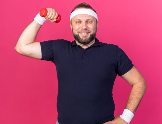 头带微笑成人斯拉夫运动男子戴头带和腕带紧张的二头肌举行哑铃隔离在粉红色的墙壁与复制空间孤立斯拉夫腕带