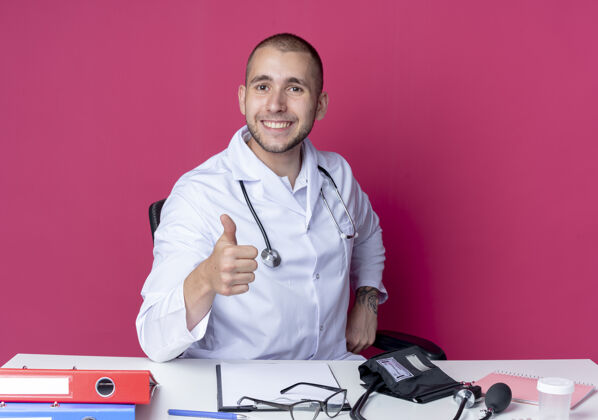 姿势面带微笑的年轻男医生穿着医用长袍 听诊器坐在办公桌旁 工作工具上的拇指被隔离在粉红色的墙上医学坐着人