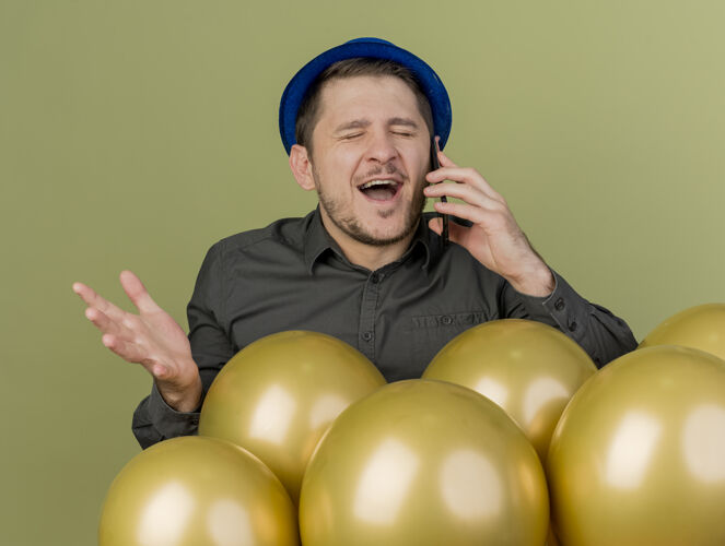 帽子戴着黑色衬衫和蓝色帽子的年轻人站在气球后面 一边打电话 一边张开橄榄绿的手 一边闭着眼睛欢喜聚会黑色欢乐
